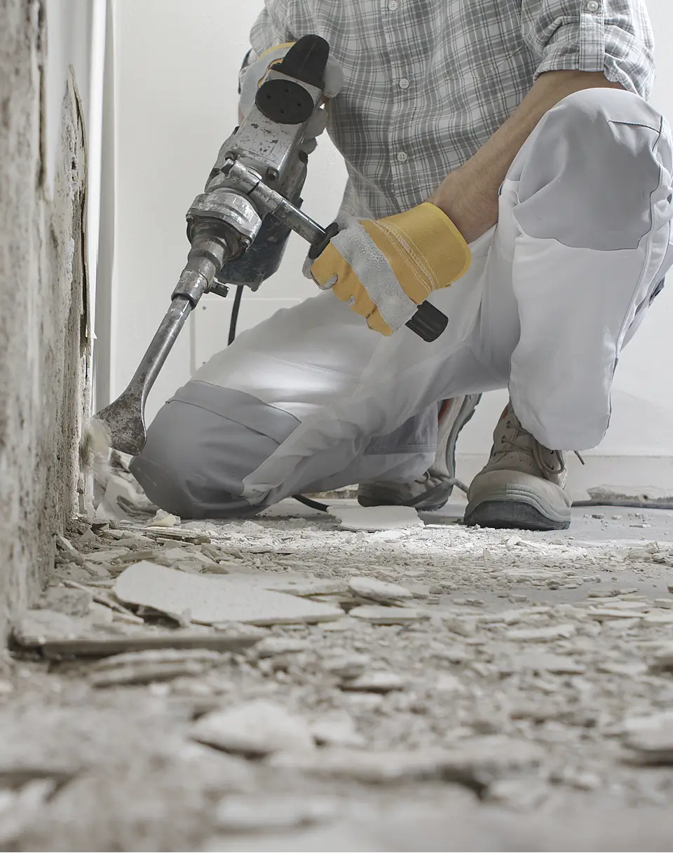 A person wearing gloves, work pants, and sturdy shoes uses a power tool to remove plaster or tiles from a wall during a home restoration. Broken debris is scattered across the dusty floor.