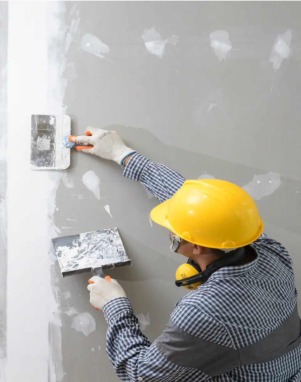 A construction worker in a yellow hard hat and gloves applies plaster to a wall with a trowel, smoothing the surface during a home restoration project in Seattle.