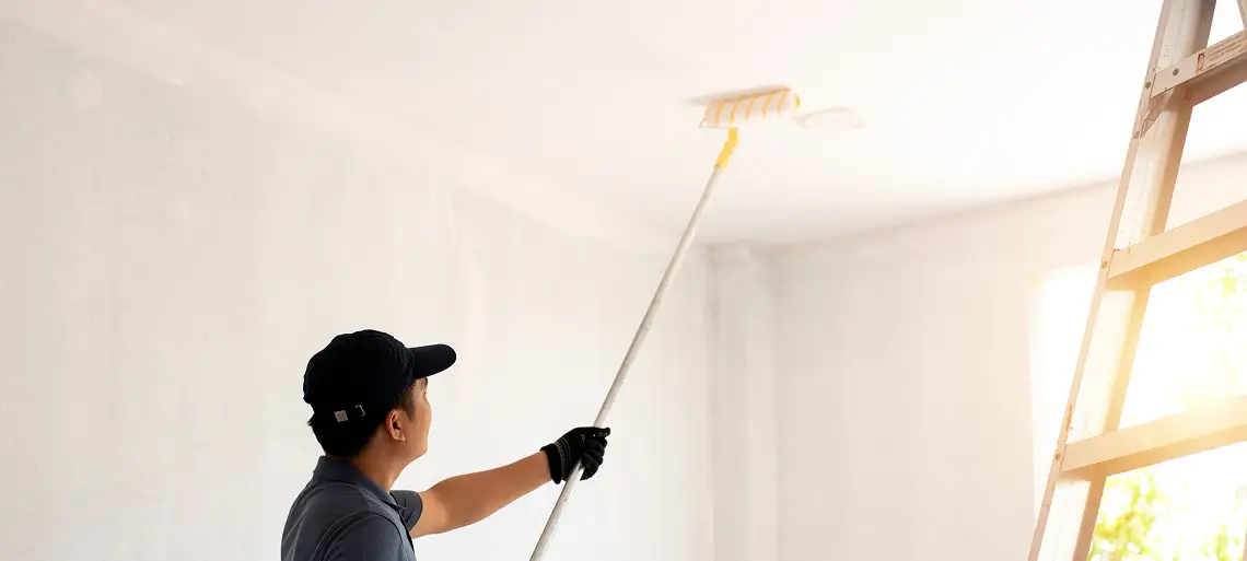 A person wearing a cap and gloves uses a paint roller with an extension pole to paint a white ceiling, part of a home restoration project. A ladder stands nearby and sunlight streams through the window, brightening the restoration Seattle scene.