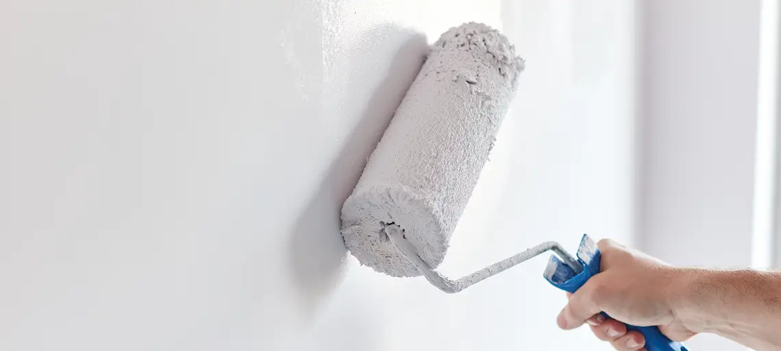 A hand holds a paint roller applying white paint to a smooth wall, with the roller covered in fresh paint—a satisfying step in home restoration.