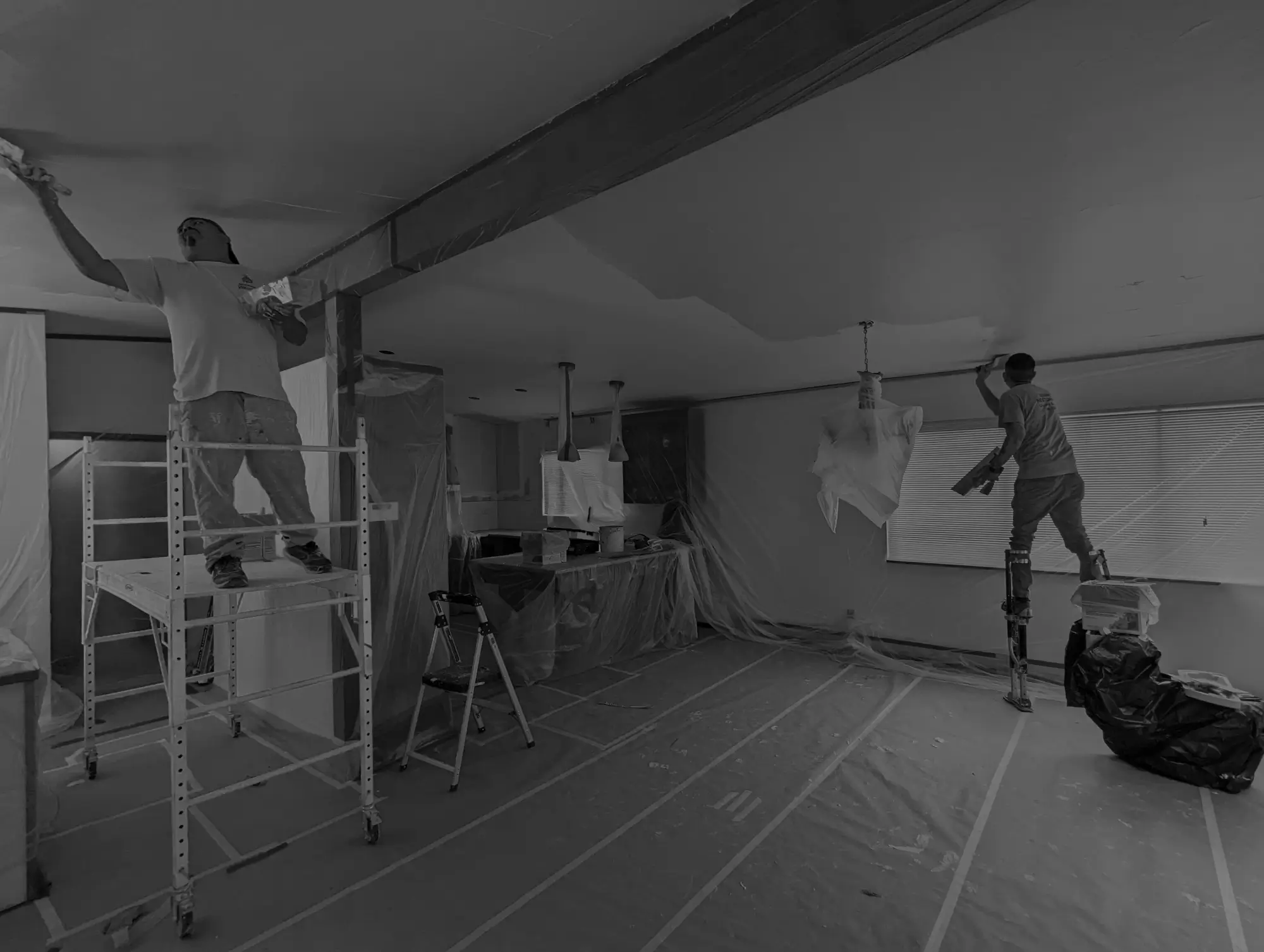 Two workers tackle a home restoration project, painting a large ceiling in a room covered with protective plastic sheets. One is on scaffolding, the other on stilts. Furniture and floors are shielded, showing careful restoration Seattle style.