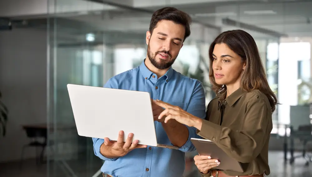 Two people standing in an office, looking at a laptop together. One person is holding the laptop and the other a tablet, both focused and engaged in discussion about home restoration projects in Seattle.