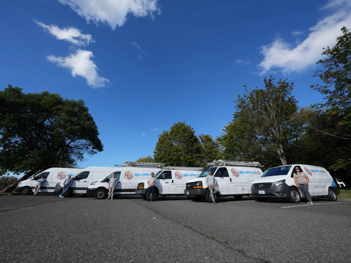 Five white service vans with company logos are parked in a row in an outdoor lot, representing restoration Seattle. Five people stand beside or lean against the vans. Trees and a blue sky with scattered clouds are visible in the background.
