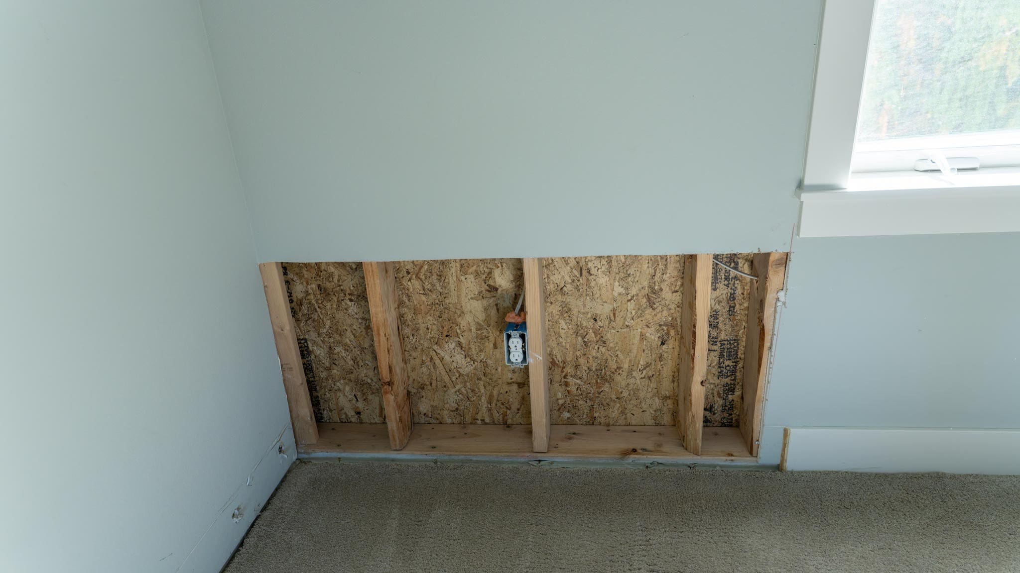 A section of drywall is removed from a wall, exposing wooden studs, insulation, and an electrical outlet—typical in home restoration projects. The area sits near a window with white trim and beige carpeted flooring.
