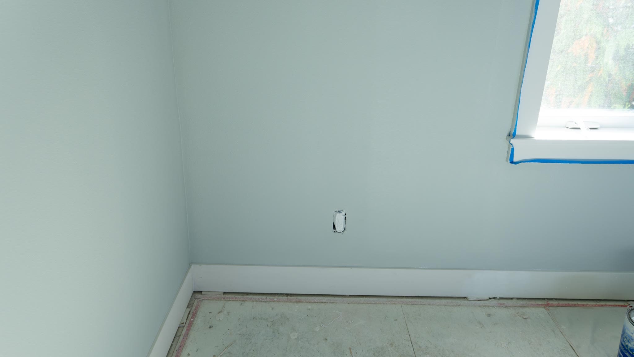 A room corner with light gray walls, a white baseboard, an unfinished floor, and a window partially covered with blue painters tape—perfect for a home restoration project. An electrical outlet without a cover is visible on the wall.