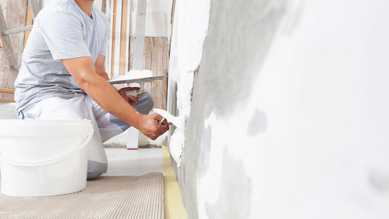 A person applies white plaster to a gray wall with a trowel during a home restoration project. A white bucket sits nearby, and various tools are visible in the background, suggesting interior renovation or repair work.