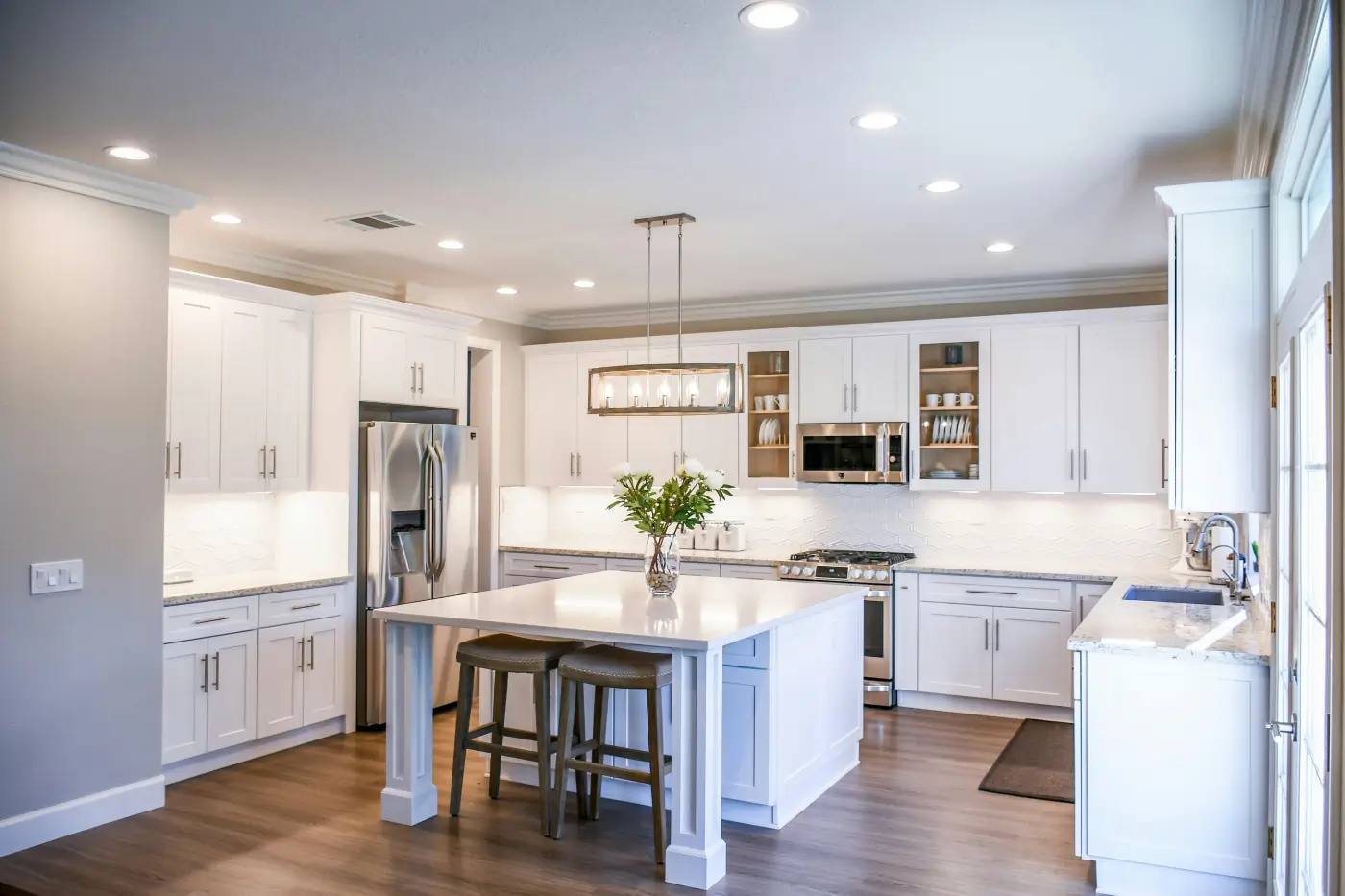 Modern kitchen with white cabinets, stainless steel appliances, a large island with two stools, wooden flooring, and pendant lighting—showcasing quality home restoration. Natural light streams in from the right onto a vase of greenery on the island.