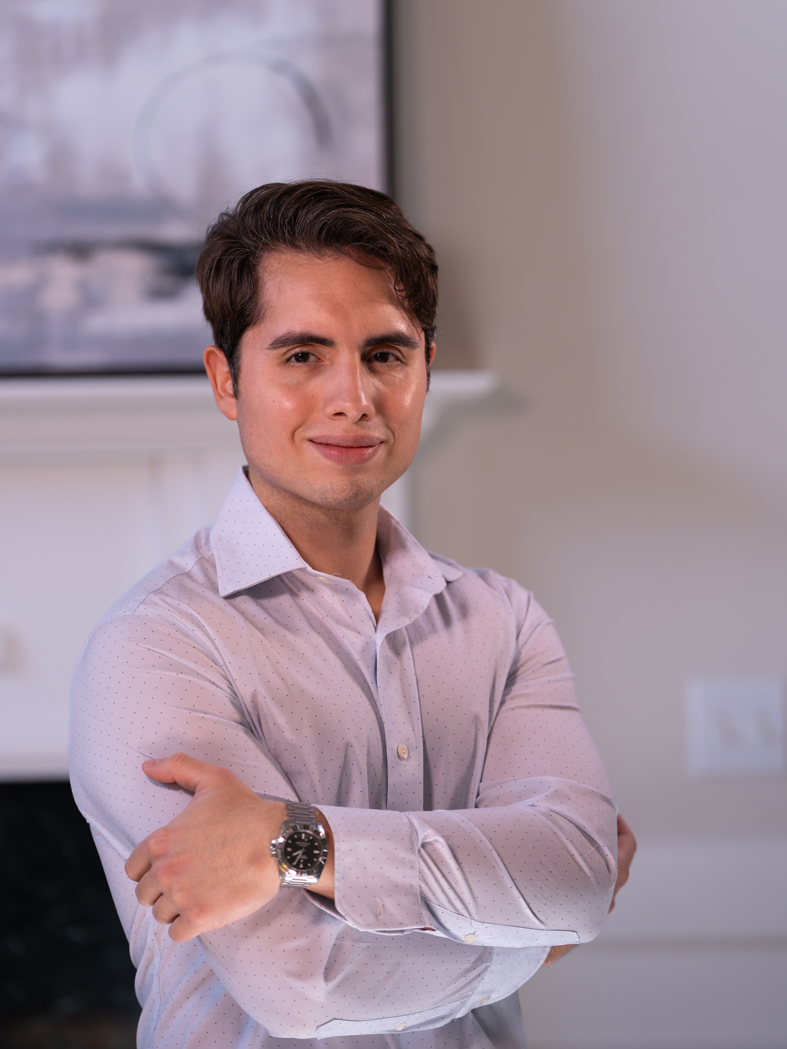 A young man with short dark hair, wearing a light-colored dress shirt and a wristwatch, stands indoors with his arms crossed, smiling confidently at the camera. A blurred painting and a fireplace are in the background.