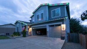 A two-story house with a double garage, gray siding, stone accents, and colorful holiday lights along the roof showcases recent home restoration. An American flag hangs near the entrance, and a car is parked inside the open garage.