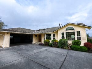 Single-story yellow house with large windows, a two-car garage, shrubs, and a wide concrete driveway under a cloudy sky—an excellent example of home restoration in Seattle.