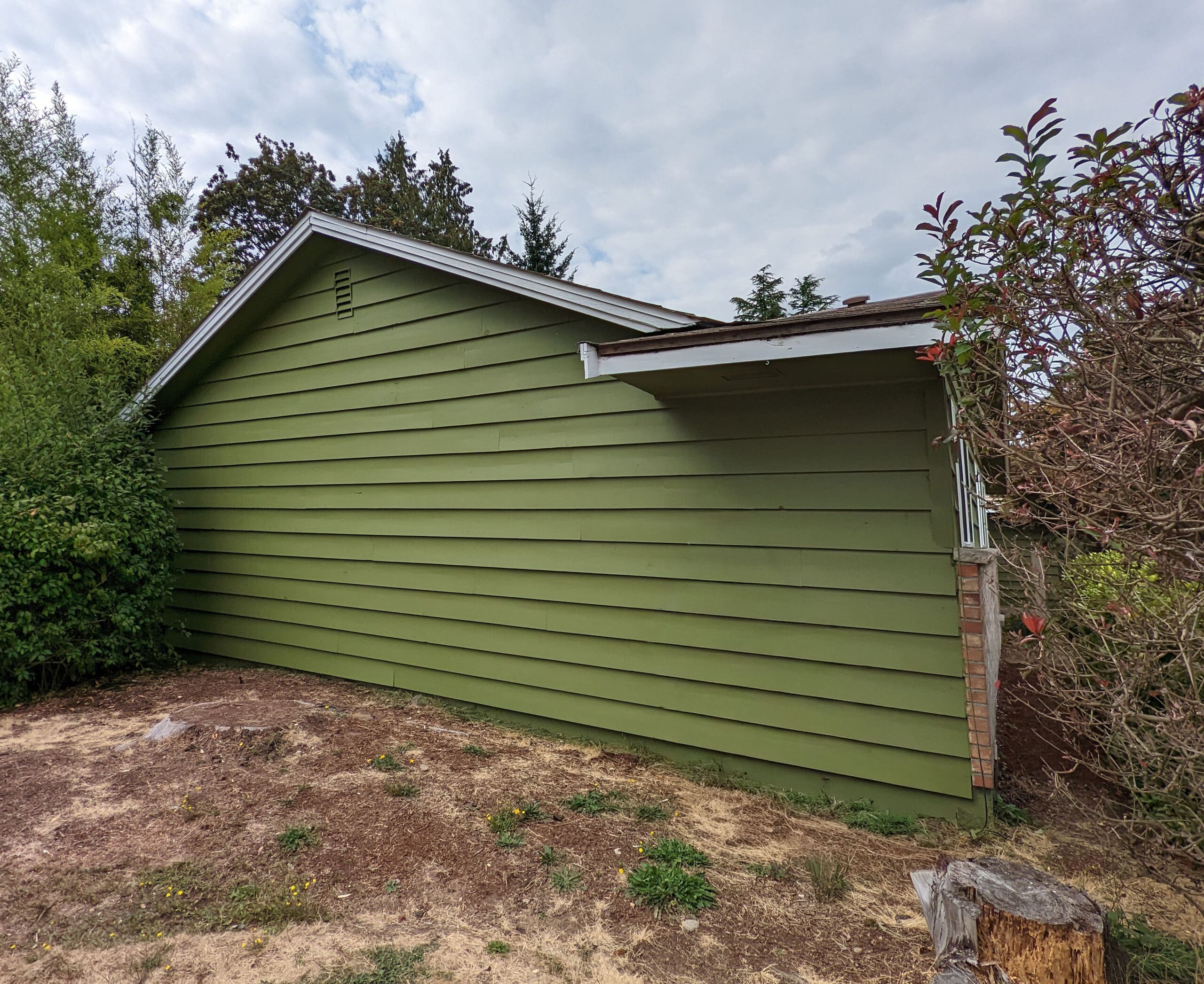 A green, single-story house with horizontal siding and a sloped roof showcases thoughtful home restoration, featuring a small covered side entrance and surrounded by sparse vegetation and trees on a cloudy day.