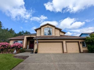 A two-story suburban house with beige siding, a double garage, and large windows. Expert home restoration in Seattle brings new life to its facade, while pink flowering bushes and a green lawn frame the front yard under a partly cloudy blue sky.