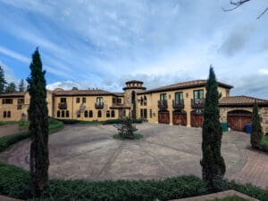 Large Mediterranean-style mansion with arched windows, balconies, stone accents, and a central circular driveway surrounded by manicured greenery—an ideal candidate for expert home restoration in Seattle. The sky above is partly cloudy.