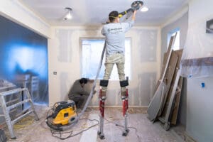 A professional drywall contractor on stilts sanding a ceiling using a dustless power sander during a home renovation.
